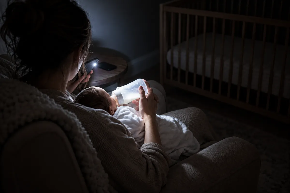 The same night feed lit by a phone flashlight: a harsh white beam on the bottle and the baby