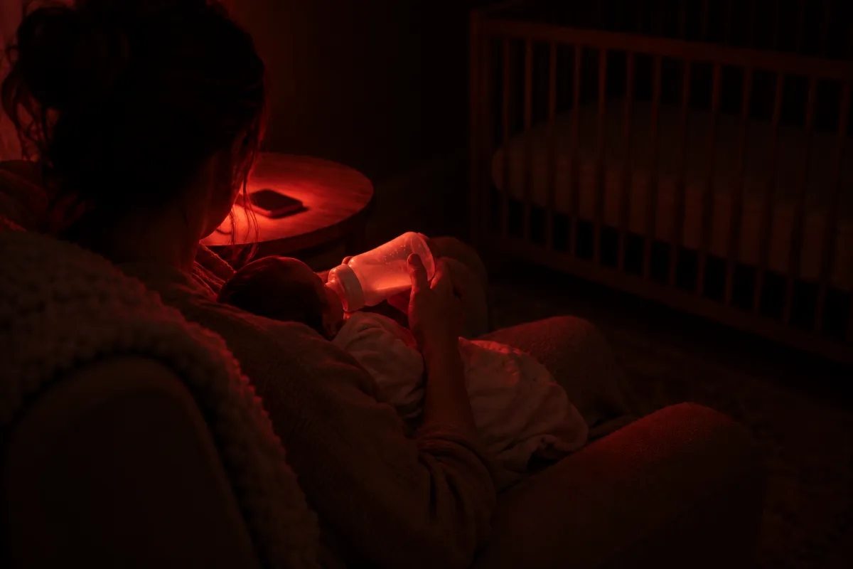A parent bottle-feeding a newborn at night by the dim red glow of a phone lying on the side table