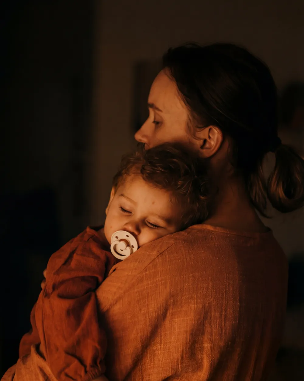 A parent holding a sleeping baby in a dim, warm-lit room