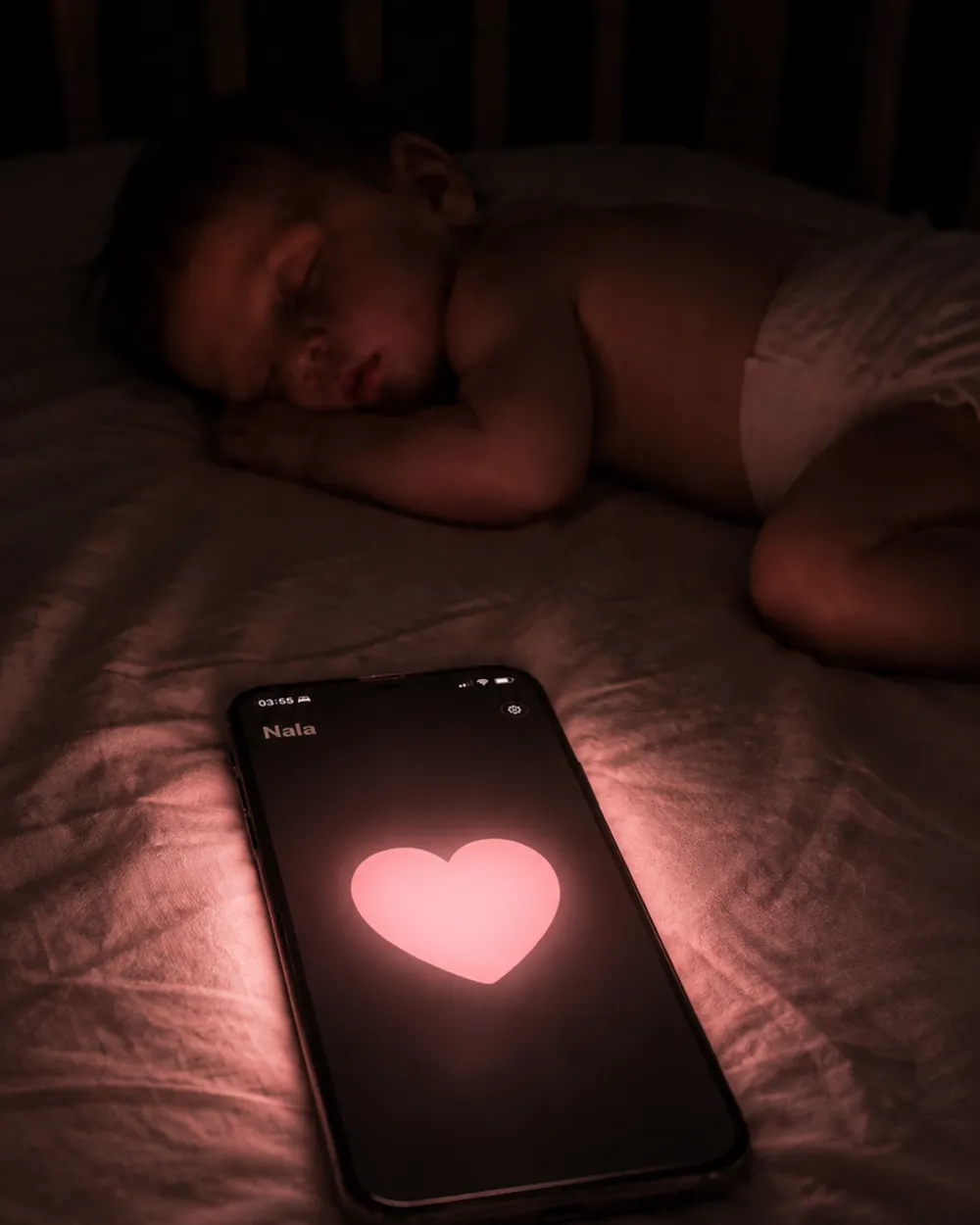A baby asleep on a bed next to an iPhone glowing with a soft pink heart